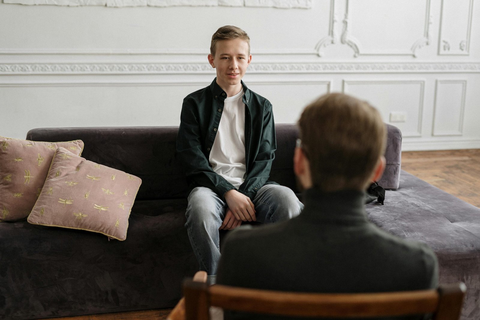 A teenage boy in a counseling session with a therapist in a modern loft setting, sitting on a sofa.